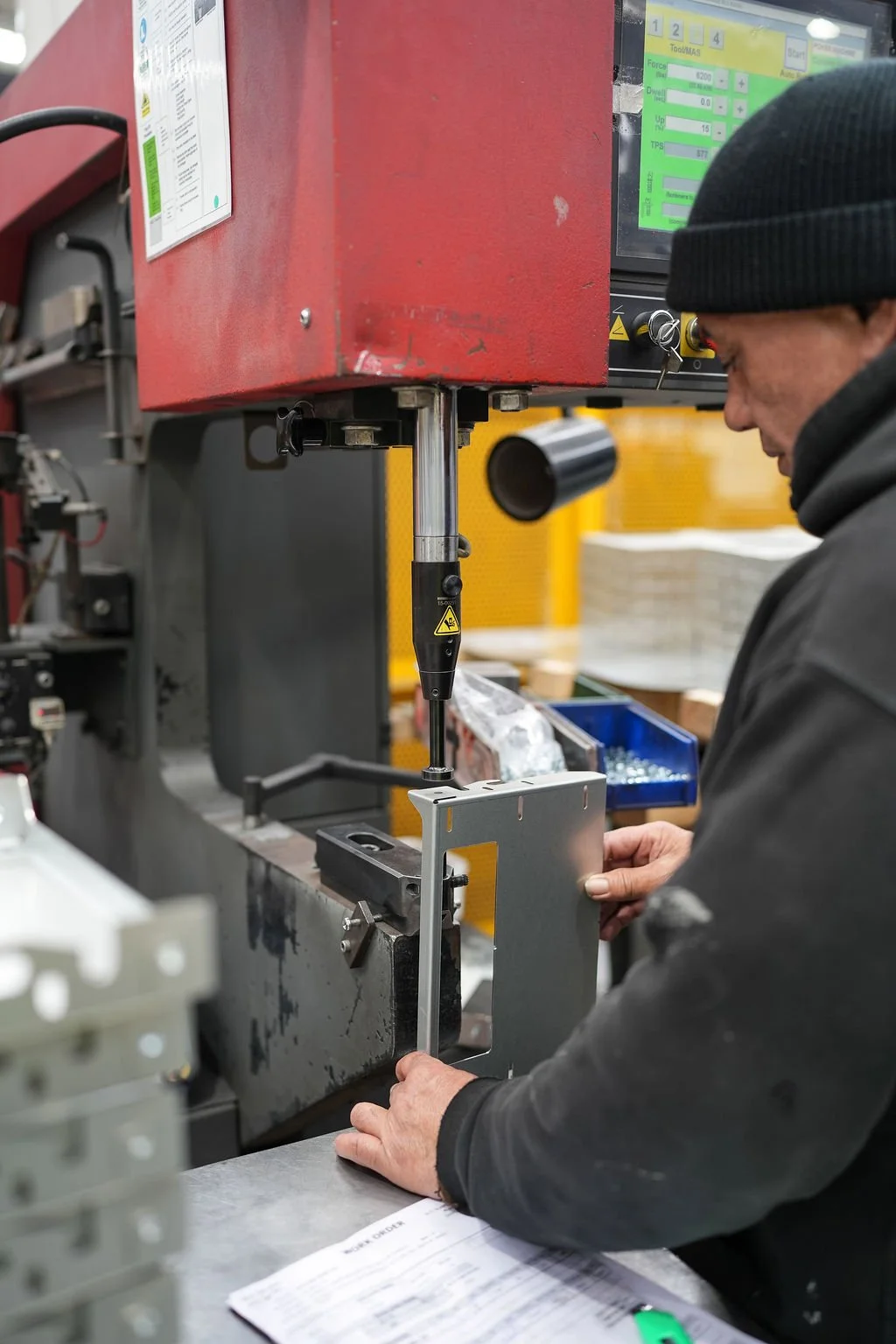 A worker in a black beanie and jacket is operating a machine drill to fasten a metal piece onto a workpiece in an industrial setting with yellow bins and a digital control panel in the background.