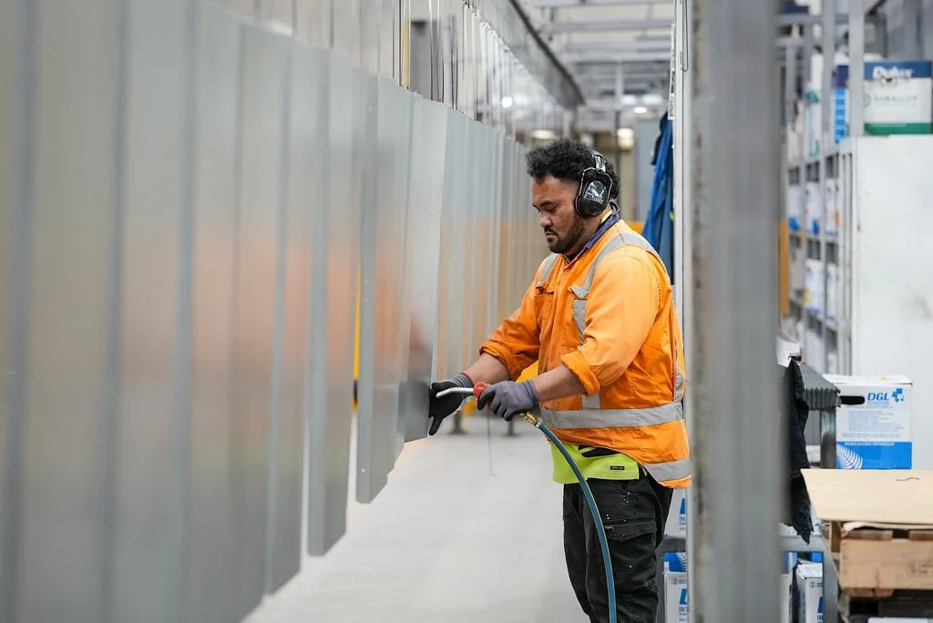 A worker in an orange safety jacket and headphones is cleaning or inspecting industrial metal panels in a warehouse.