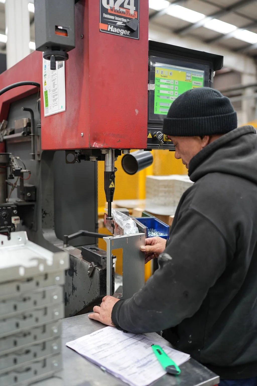A worker wearing a black beanie and hoodie operating a piece of industrial machinery in a factory setting.