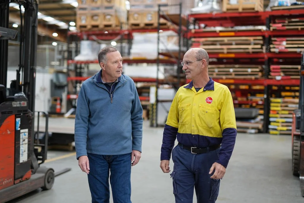 Two men talking in a warehouse, one wearing a blue jacket and jeans, the other in a yellow and navy uniform, with shelves of wood planks in the background.