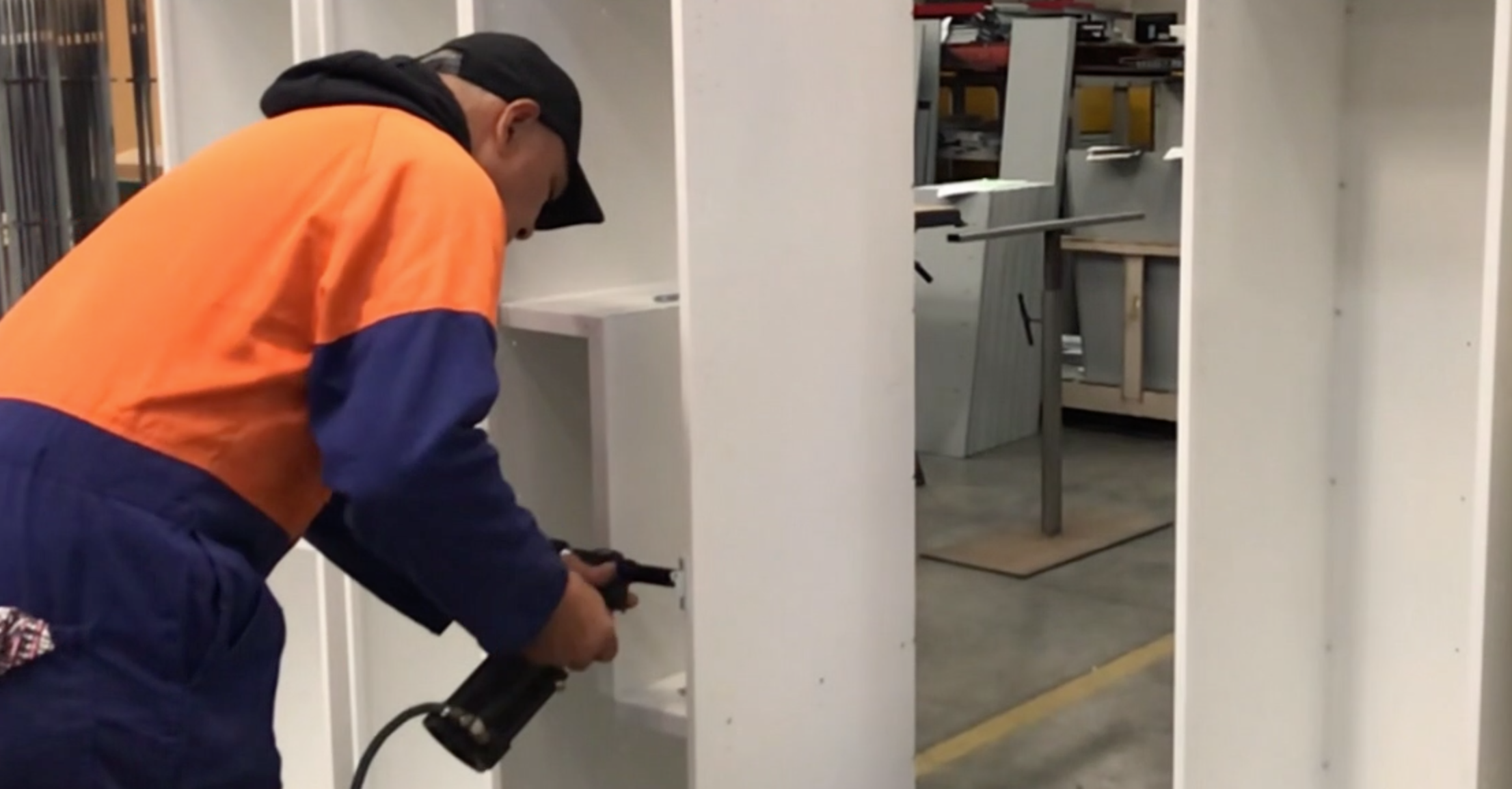 A man wearing an orange and navy blue jacket, a black cap, and glasses, is using a power drill on a white wall in a building under construction or renovation.