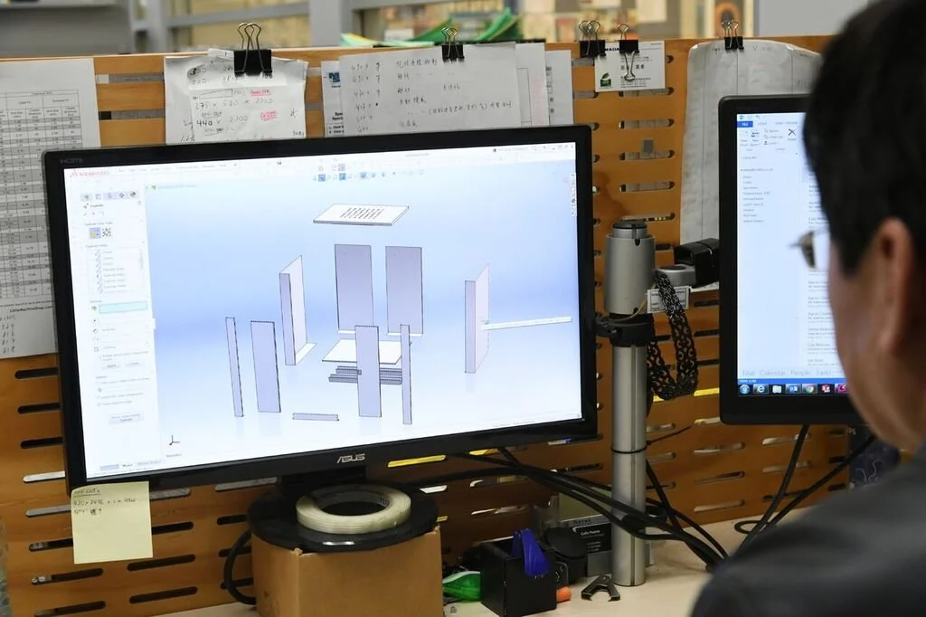 A person working on a computer with CAD software displaying a 3D model of a structure made of multiple upright panels. The workspace has various papers clipped to a wooden board and other office supplies around.