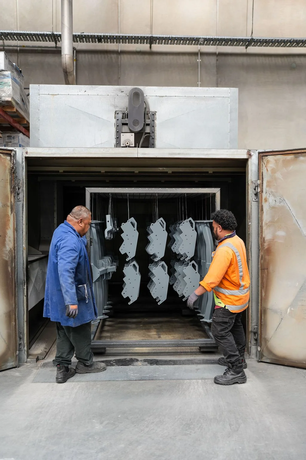 Two workers inspect gray metal parts hanging inside an industrial oven or curing chamber in a factory setting.