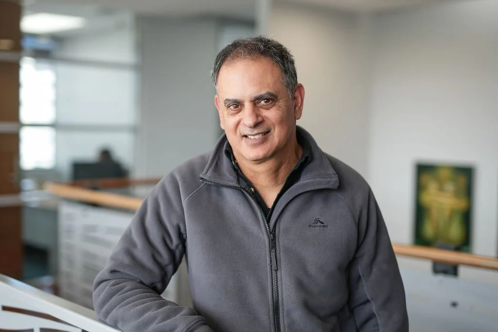A middle-aged man with dark hair and a friendly expression, wearing a gray zip-up jacket, standing indoors in a modern building.