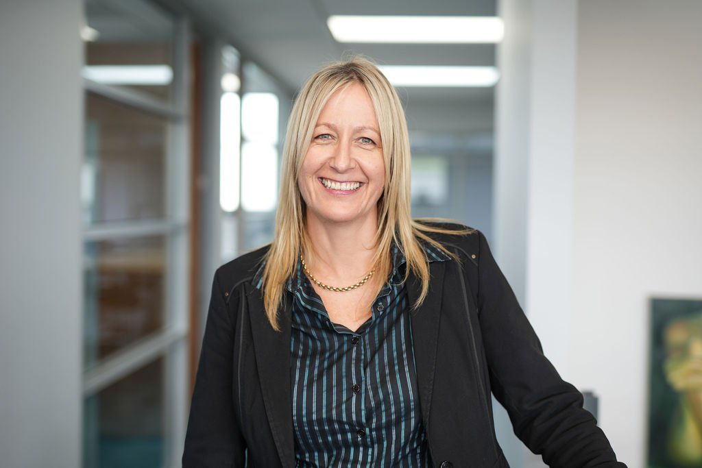 A smiling woman with blonde hair wearing a black blazer and blue striped shirt in an indoor office setting.