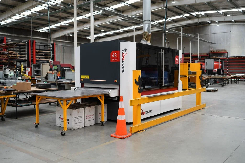 Large industrial machine in a warehouse or factory, surrounded by worktables and shelving units, with a safety cone and guardrail around it.