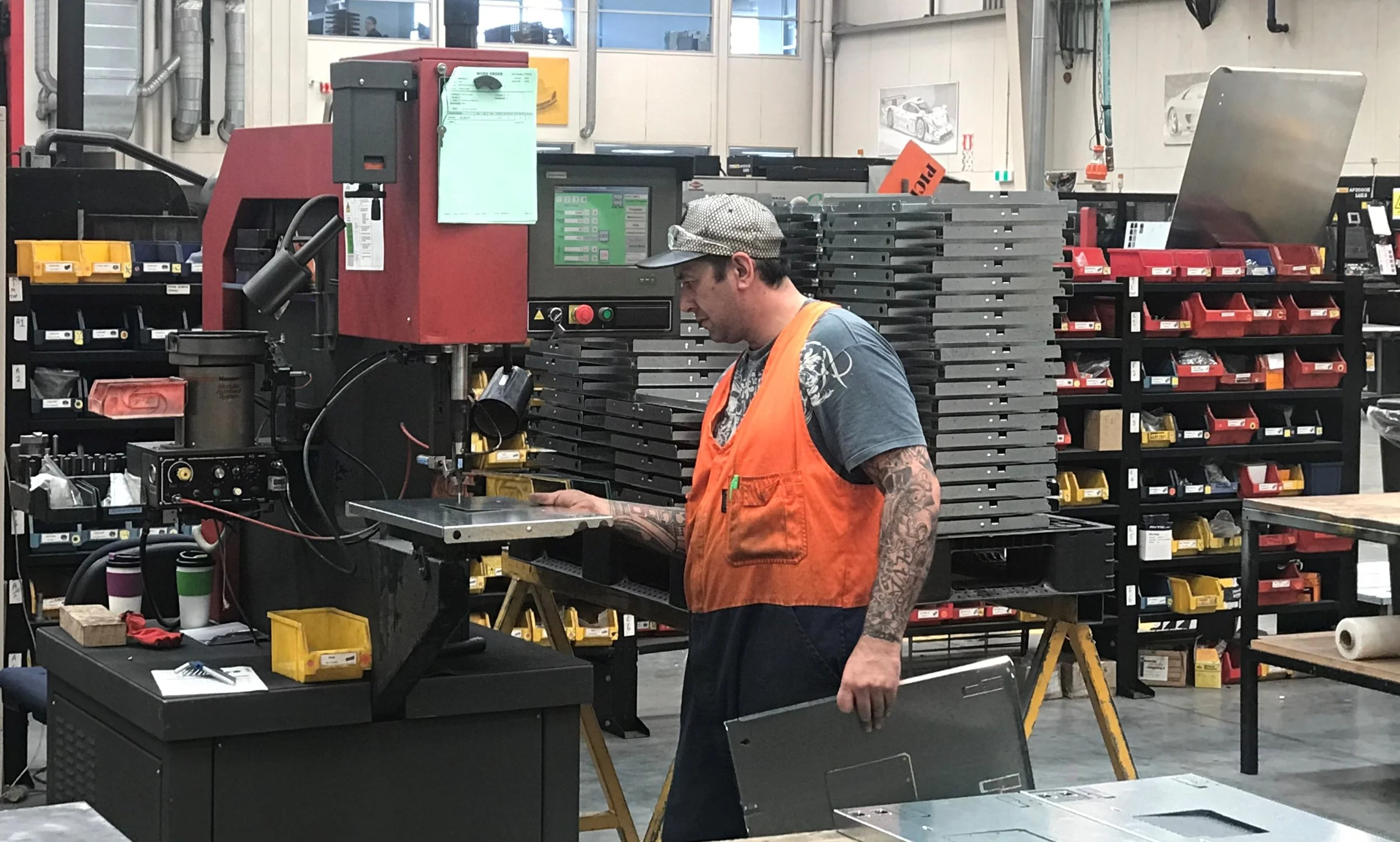 A man in an orange safety vest and a gray cap working at a machine in an industrial environment with tools and parts on shelves around him.