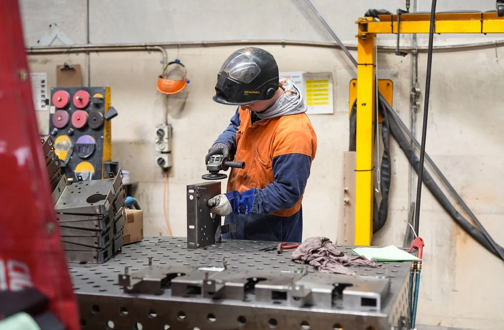 A worker wearing safety gear, including a helmet and gloves, is working with metal parts in a workshop or industrial setting.