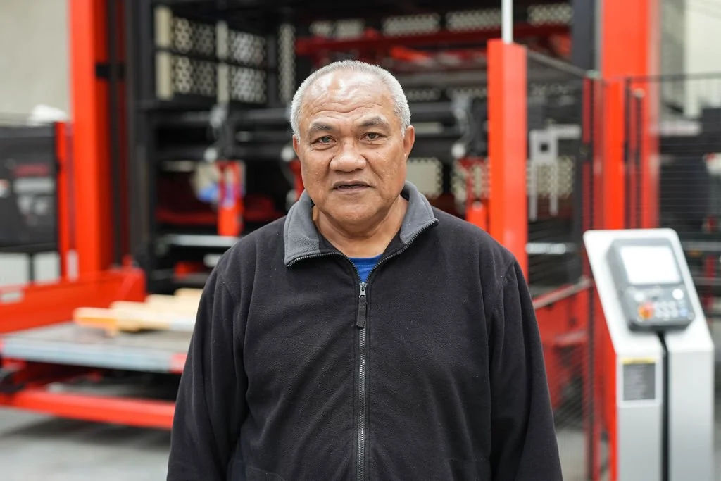 An older man with gray hair standing in a warehouse or industrial setting, with red shelving and some equipment in the background.