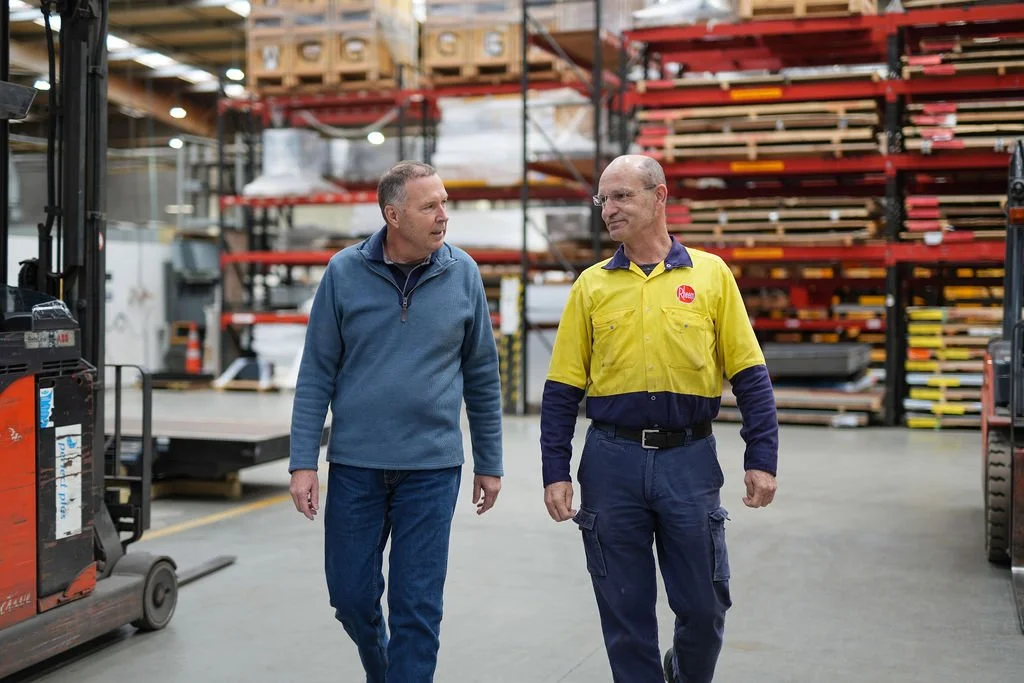 Two men walking and talking inside a hardware store or warehouse with shelves stocked with building materials and wood planks in the background.