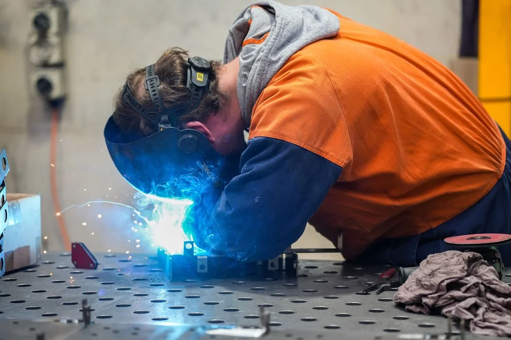 A person welding at a workbench, wearing safety goggles and a gray and orange jacket, with bright sparks and blue light from the welding process.