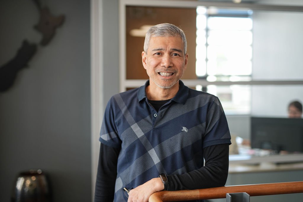 A middle-aged man with gray hair and a beard, smiling, standing in an office environment with cubicles and people working in the background.
