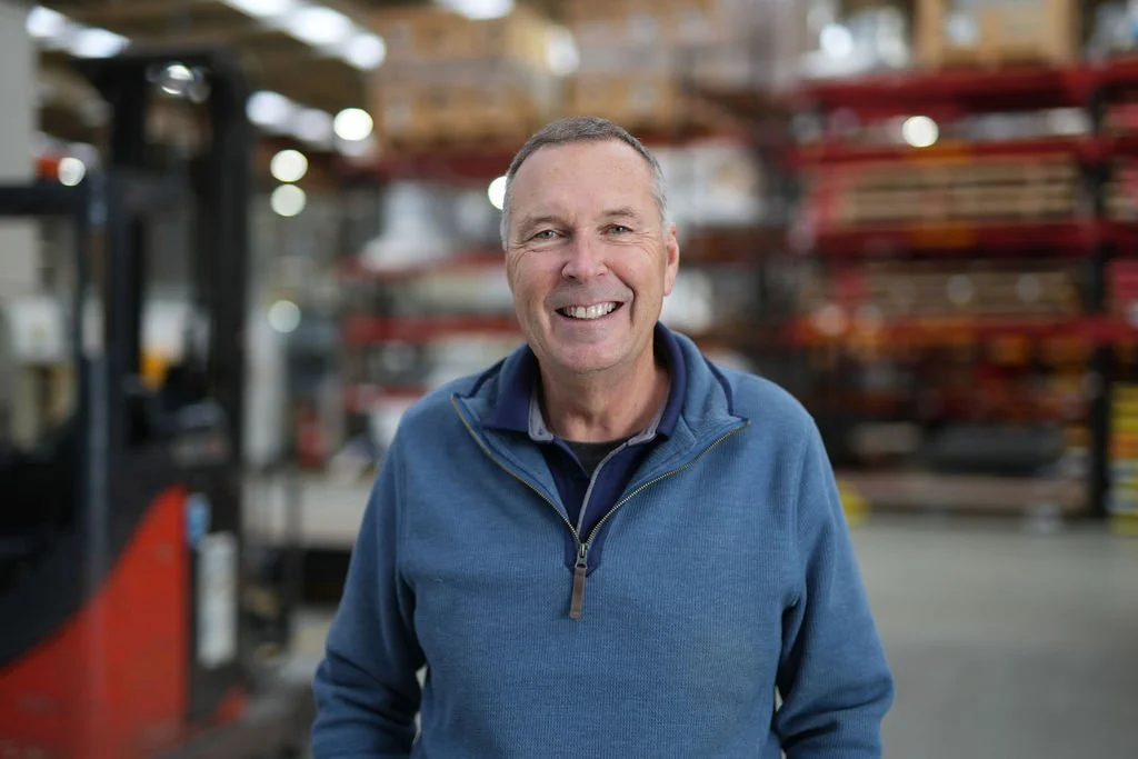 A smiling middle-aged man wearing a blue jacket standing inside a warehouse with shelves of products in the background.