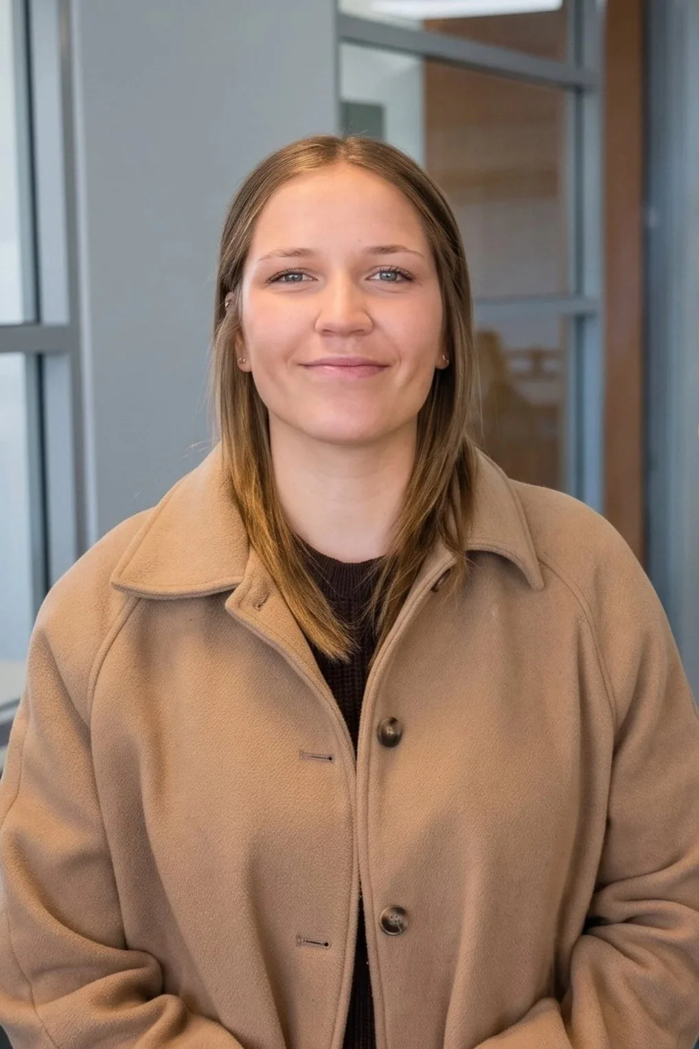 A young woman with shoulder-length light brown hair smiling, wearing a tan coat, standing indoors near large windows.