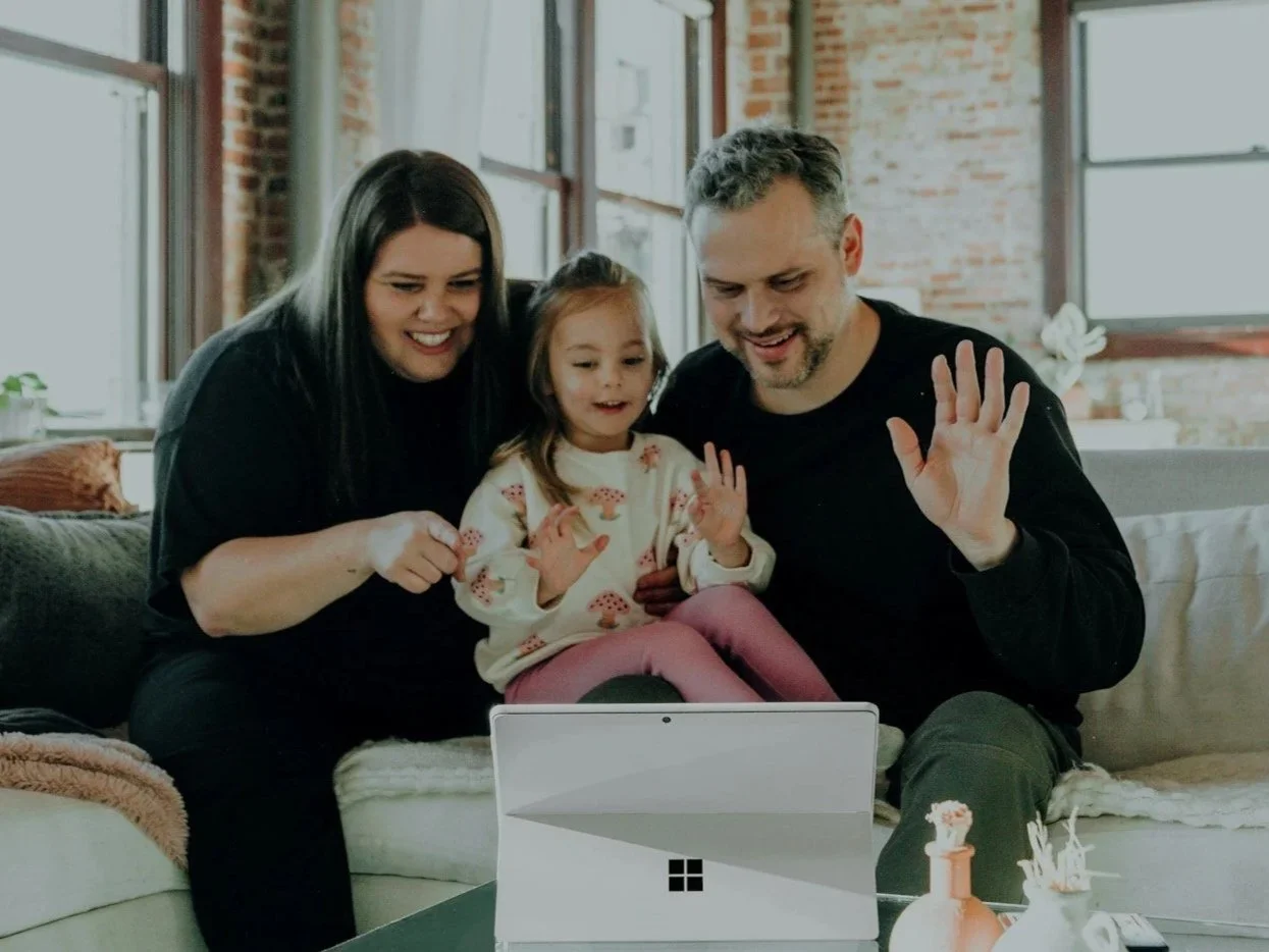 Family of three, including a woman, a man, and a young girl, sitting on a couch and waving at a tablet on a table, smiling and engaging in a video call in a bright living room with windows and brick walls.
