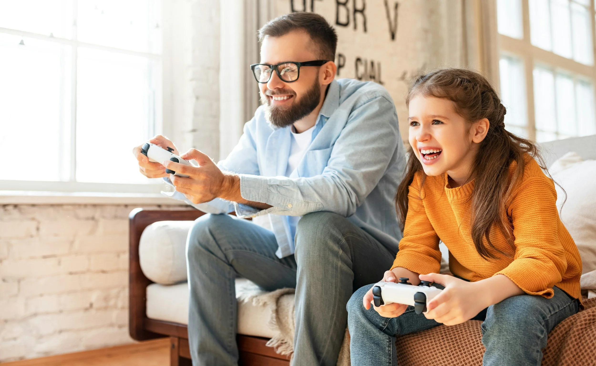 A smiling man with glasses and a beard, wearing a light blue shirt, sits on a couch playing a video game with a young girl with long hair, wearing an orange sweater, who also has a game controller in her hands. They are in a bright room with large windows.