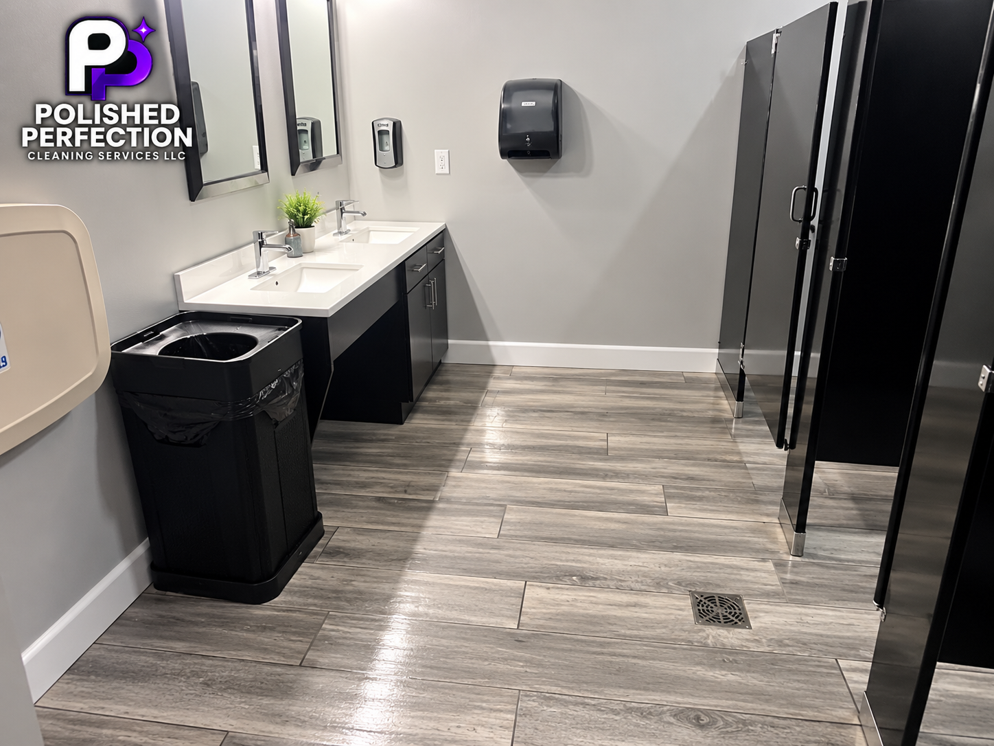 A clean restroom with two sinks, a mirror, a soap dispenser, a paper towel dispenser, a trash bin, and black lockers. There is a small plant on the sink counter and a floor drain in the wooden floor.