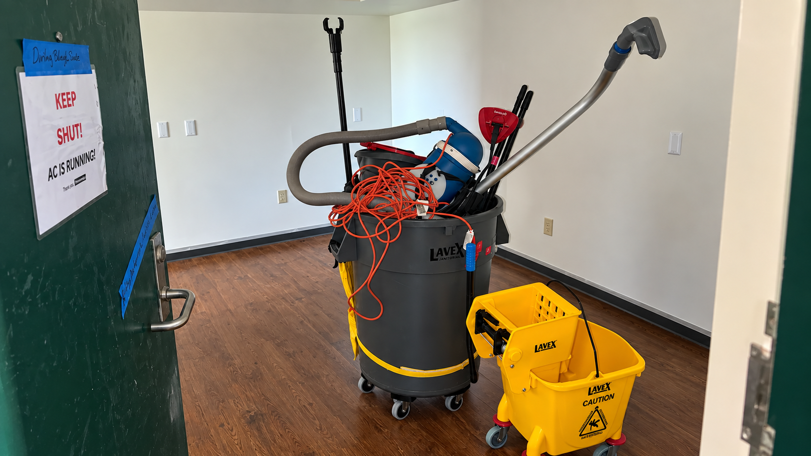 A cleaning closet containing a large black and yellow wet vacuum with orange extension cords, a red dustpan, black broom, and other cleaning supplies; a yellow mop bucket and caddy labeled 'Lavex Caution' with cleaning tools, on a wooden floor in a room with white walls.