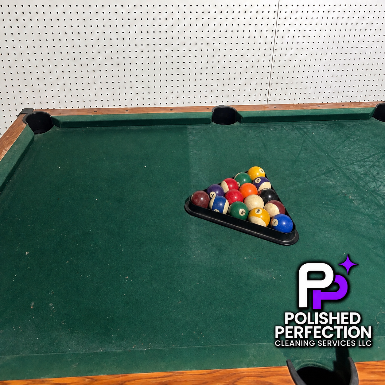 A pool table with a rack of billiard balls arranged in a triangle on the green felt surface, with a white pegboard wall in the background.