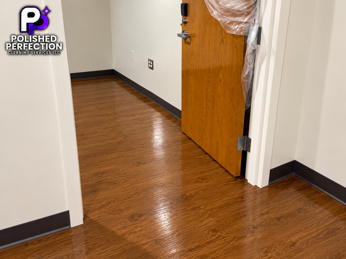 Office room with shiny wood flooring, white walls, black baseboards, and a partially open wooden door with plastic wrapped around the edge.