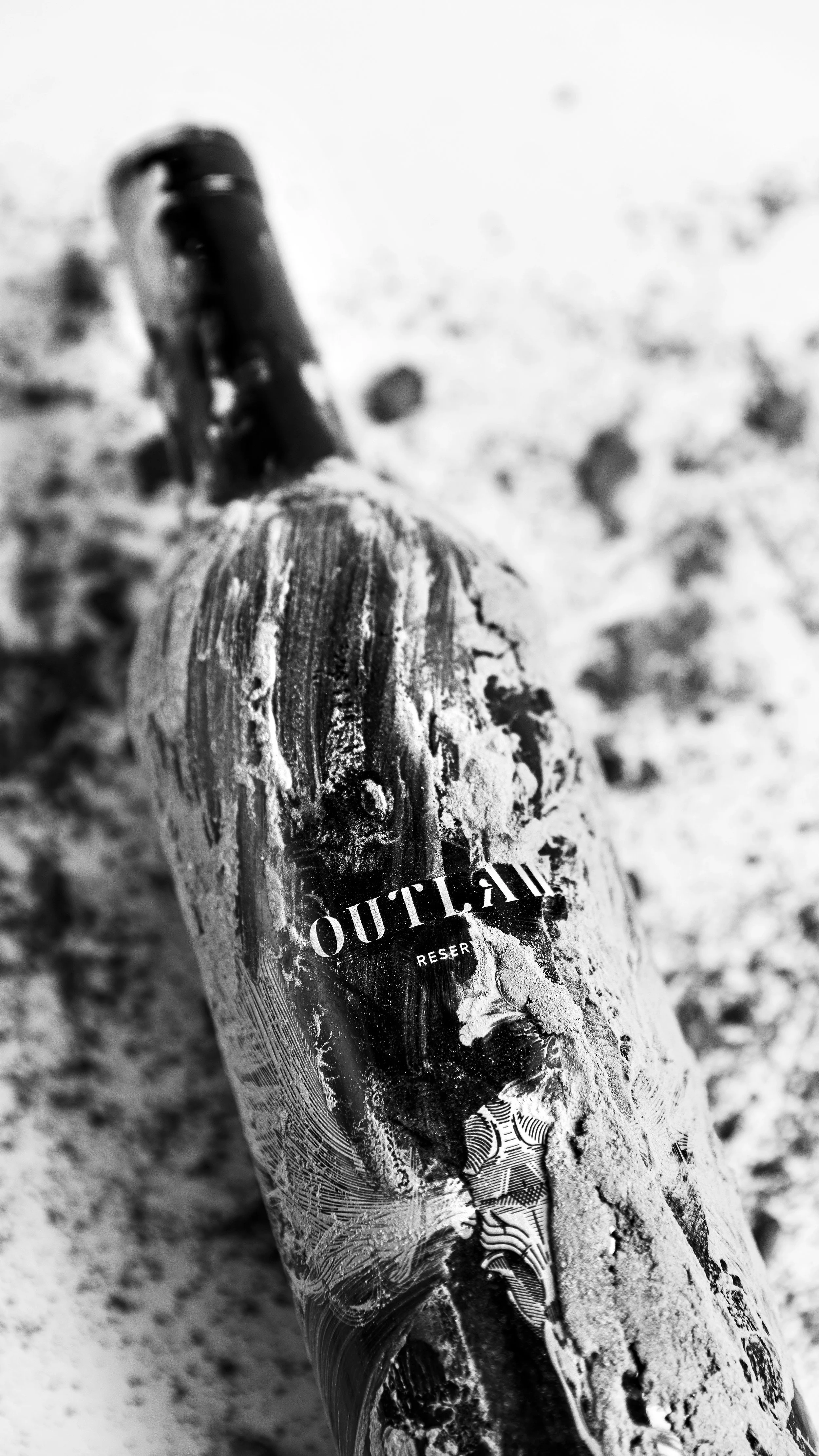 A discarded, weathered plastic bottle with the label 'OUTLAW' partially obscured, lying on sandy ground.