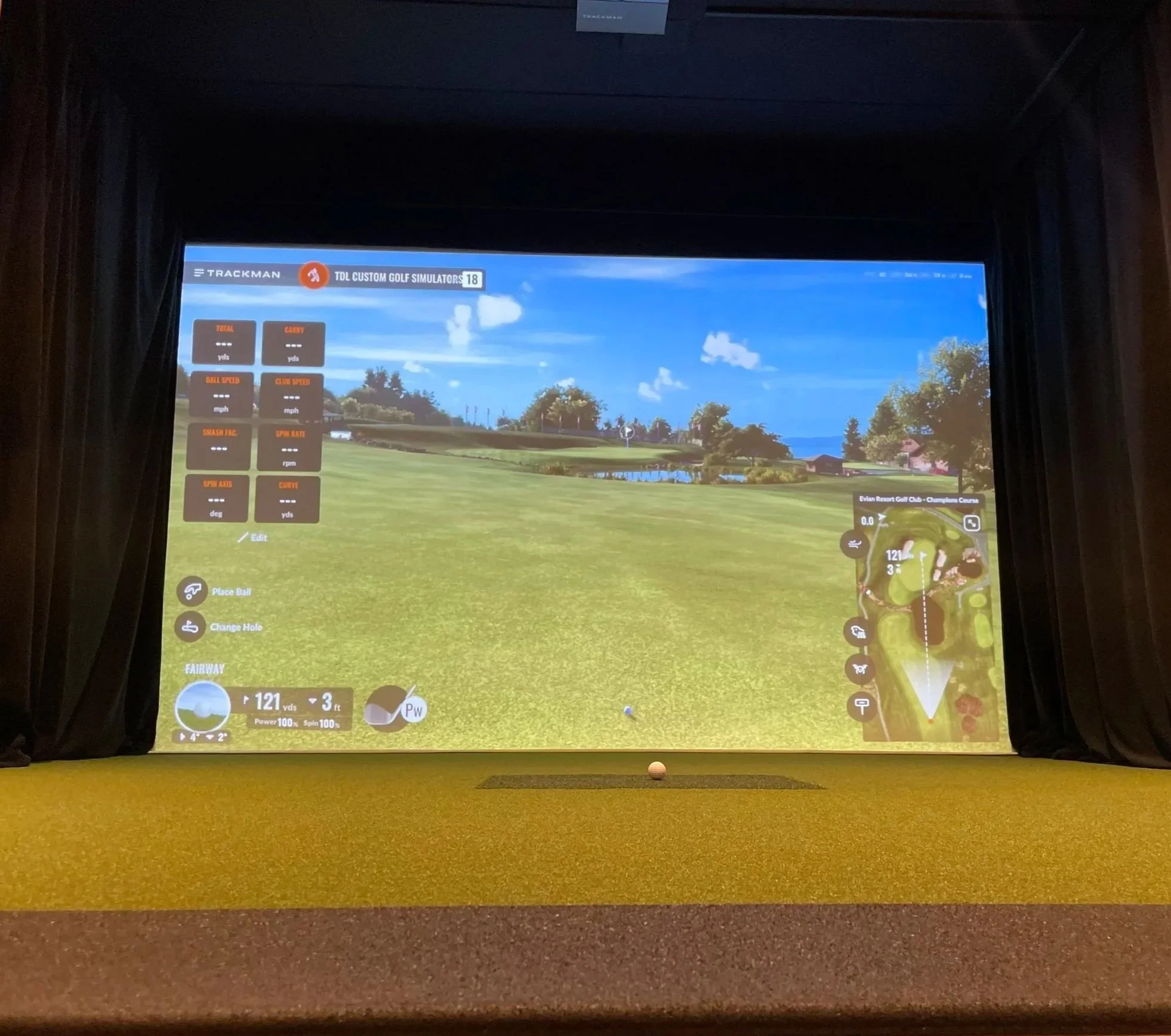 Indoor golf simulator with a large screen displaying a virtual golf course, showing a tee shot ready with the golf ball placed on a tee on the floor.