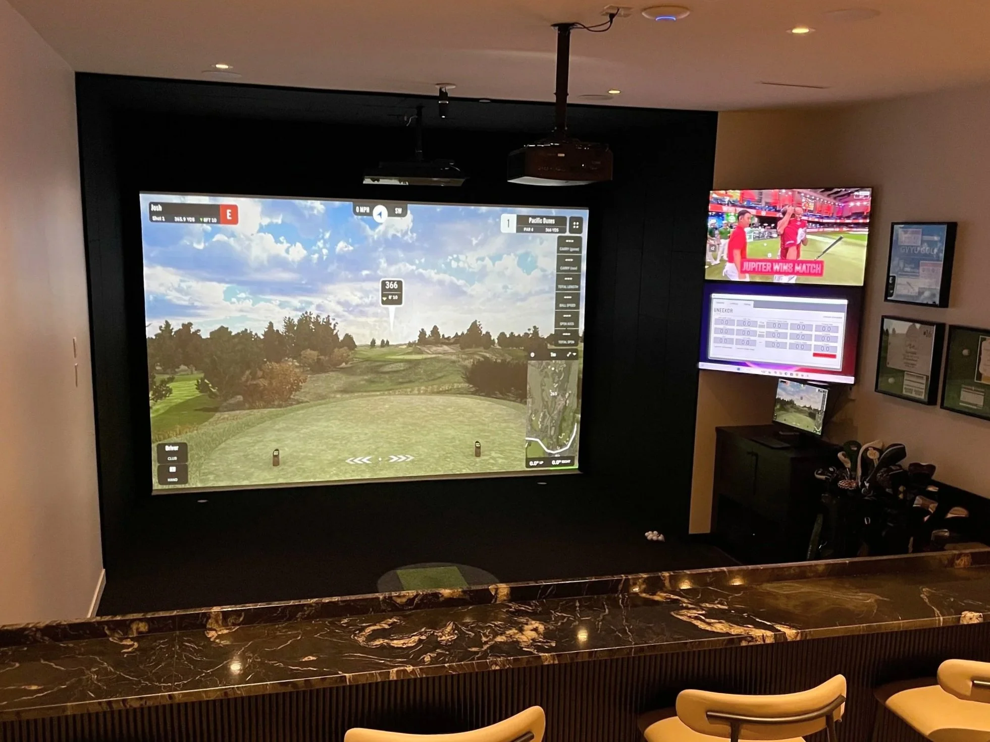 Indoor golf simulation room with large screen displaying a golf course, multiple smaller screens showing sports and game info, and seating area with chairs and a marble counter in the foreground.