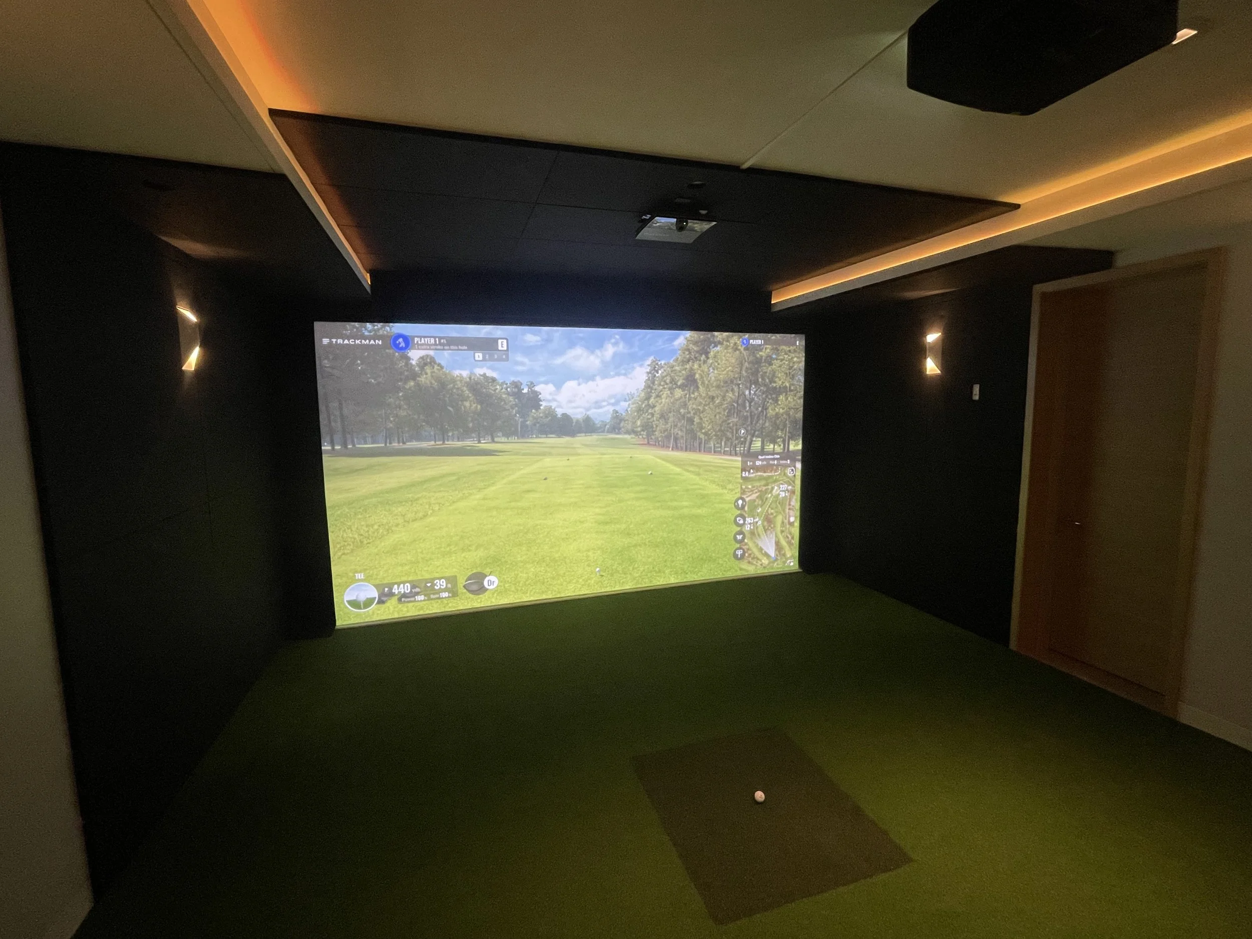 Indoor golf simulator room with a large screen displaying a golf course view, green turf flooring, and a golf ball on a small mat.