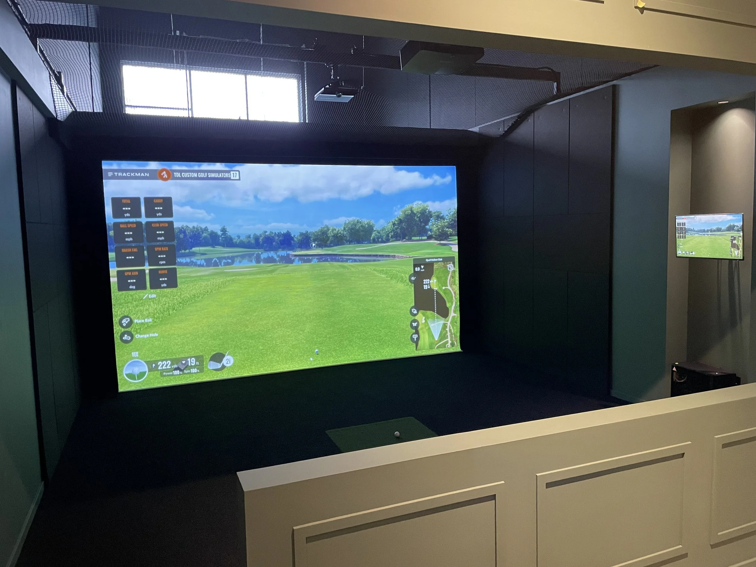 Indoor golf simulator room with a large screen showing a virtual golf course, control options on the left, and a golf ball on a tee in front of the screen.
