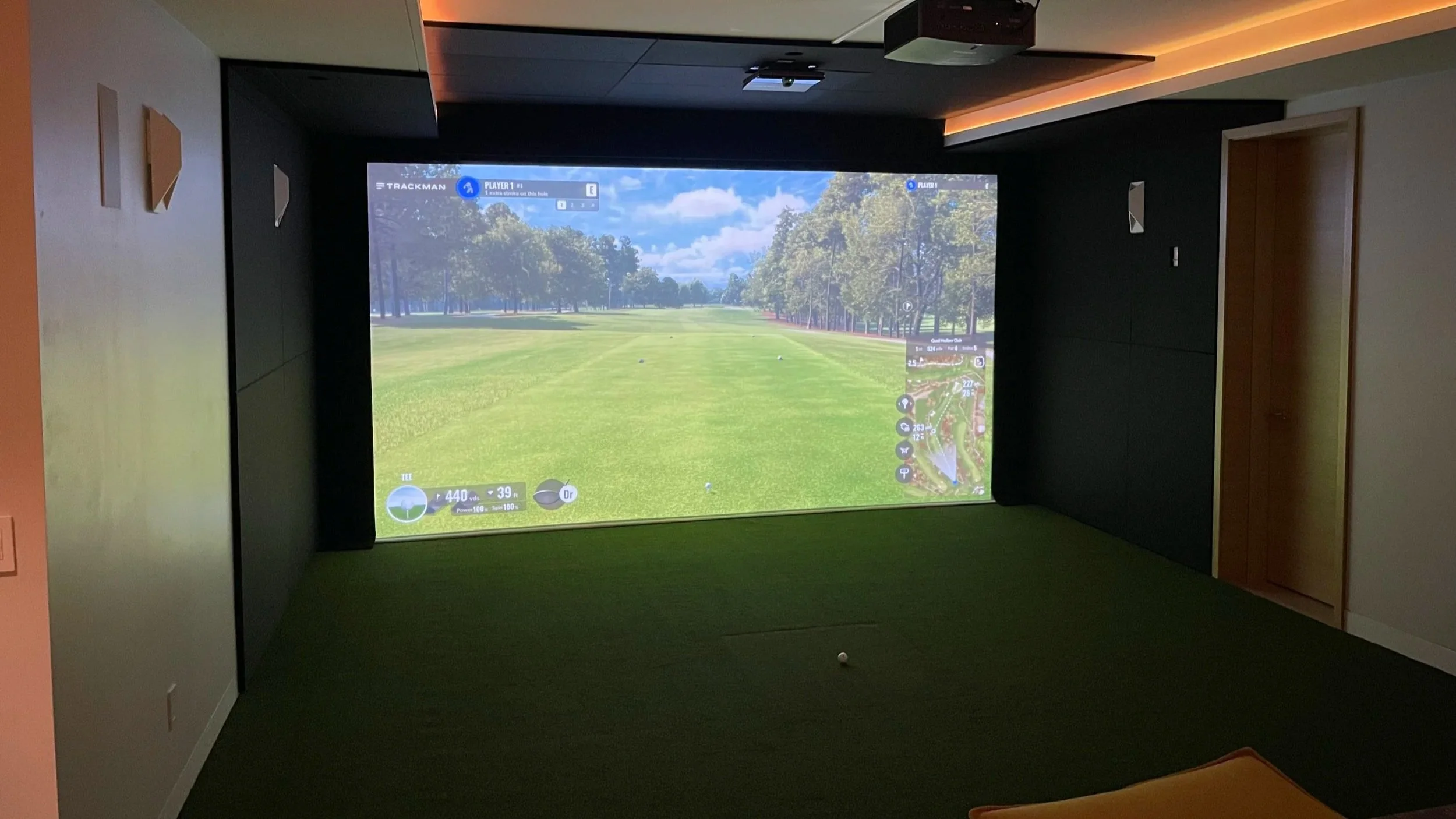 Indoor golf simulator room with a large screen showing a golf course, a small golf ball on artificial turf, and a seating area with an orange cushion.