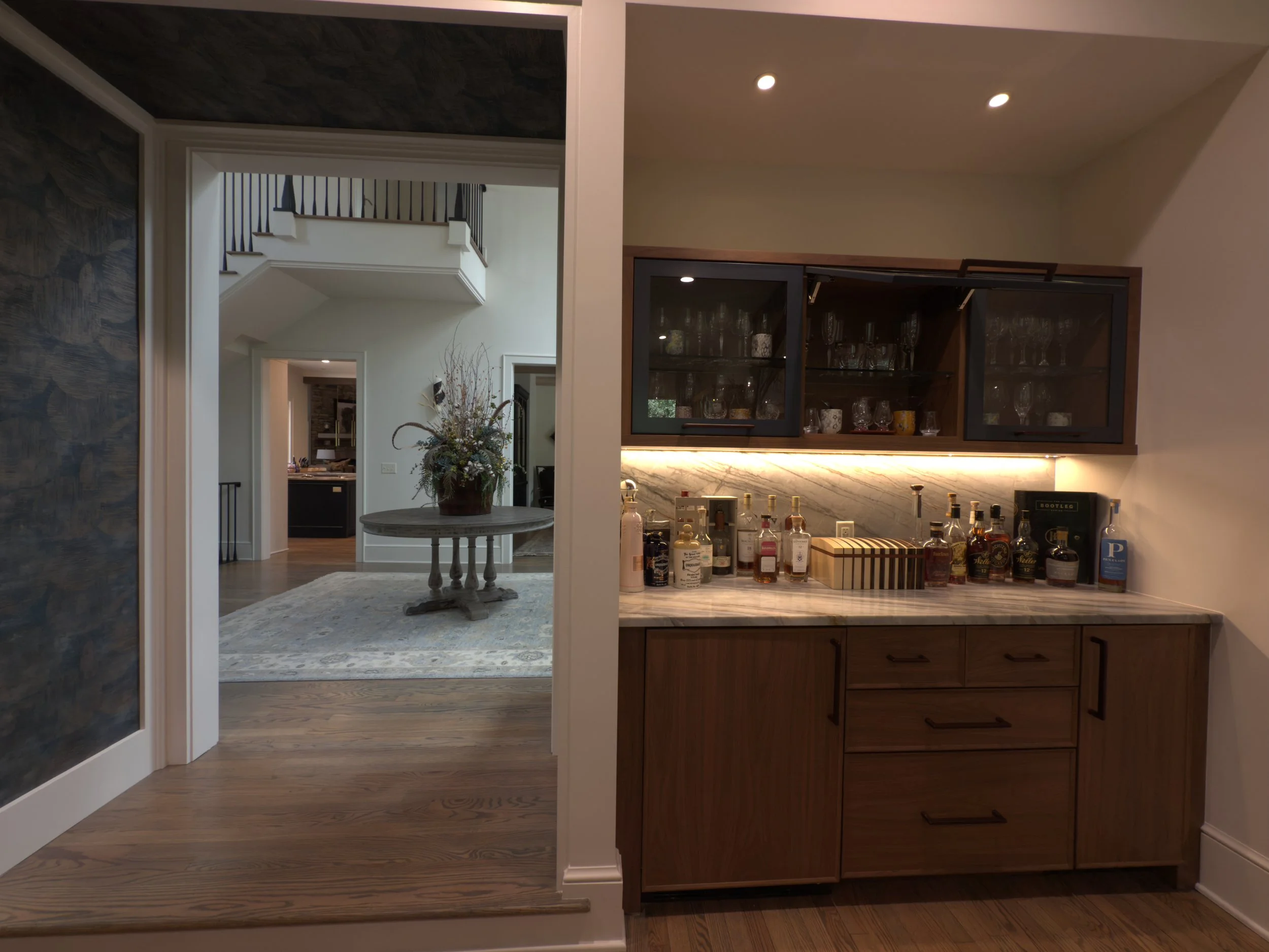 Home bar with a marble countertop, various bottles of alcohol, glassware, and wooden cabinets with glass doors above. Recessed lighting includes two round lights in the ceiling.