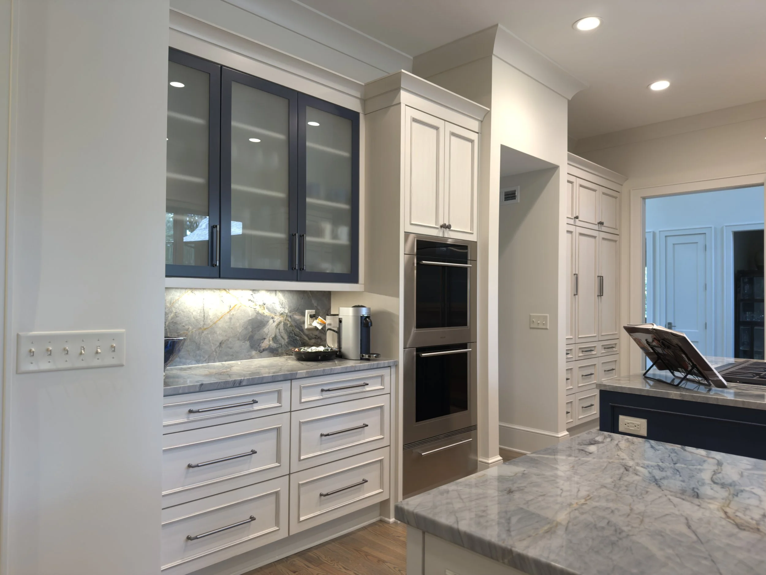 Modern kitchen with white cabinetry, marble countertops, blue glass-front upper cabinets, built-in oven, and a coffee maker on the counter.