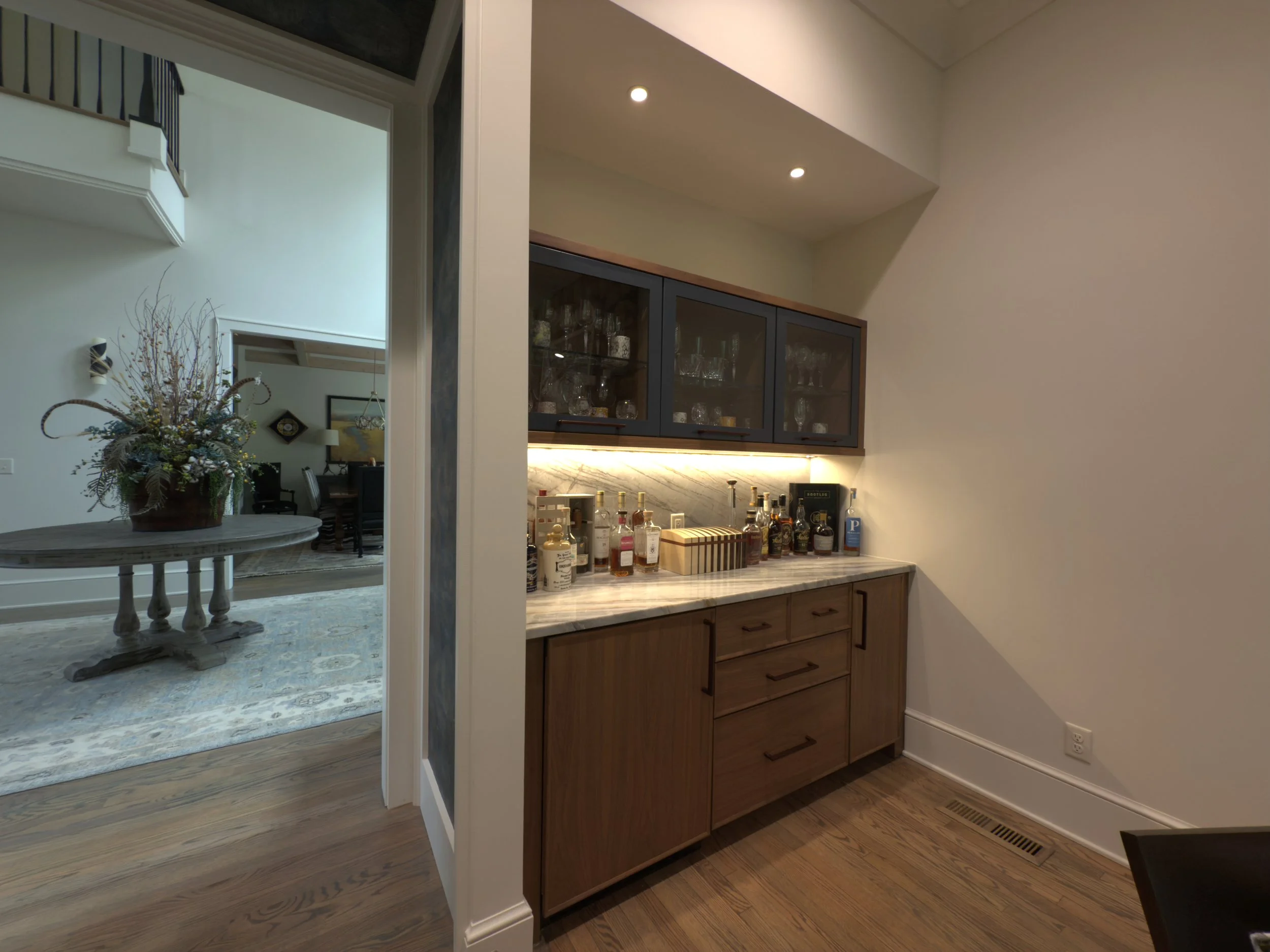 Home bar area with a granite counter, glass cabinetry, and bottles of alcohol, with adjacent dining room visible in the background.