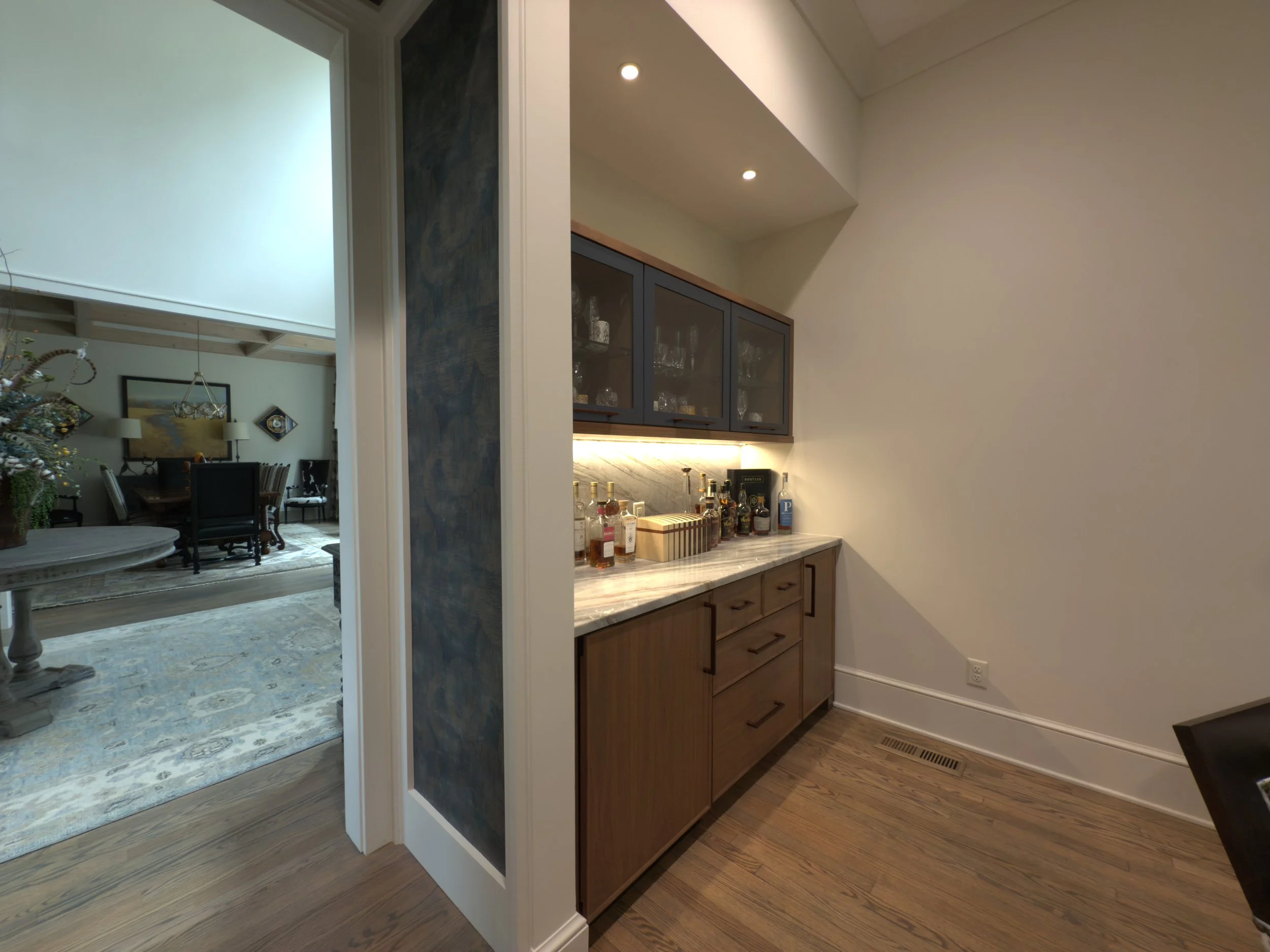 Home bar area with a marble countertop, wooden cabinets, glass-fronted wall cabinets, and various bottles of alcohol under warm lighting.