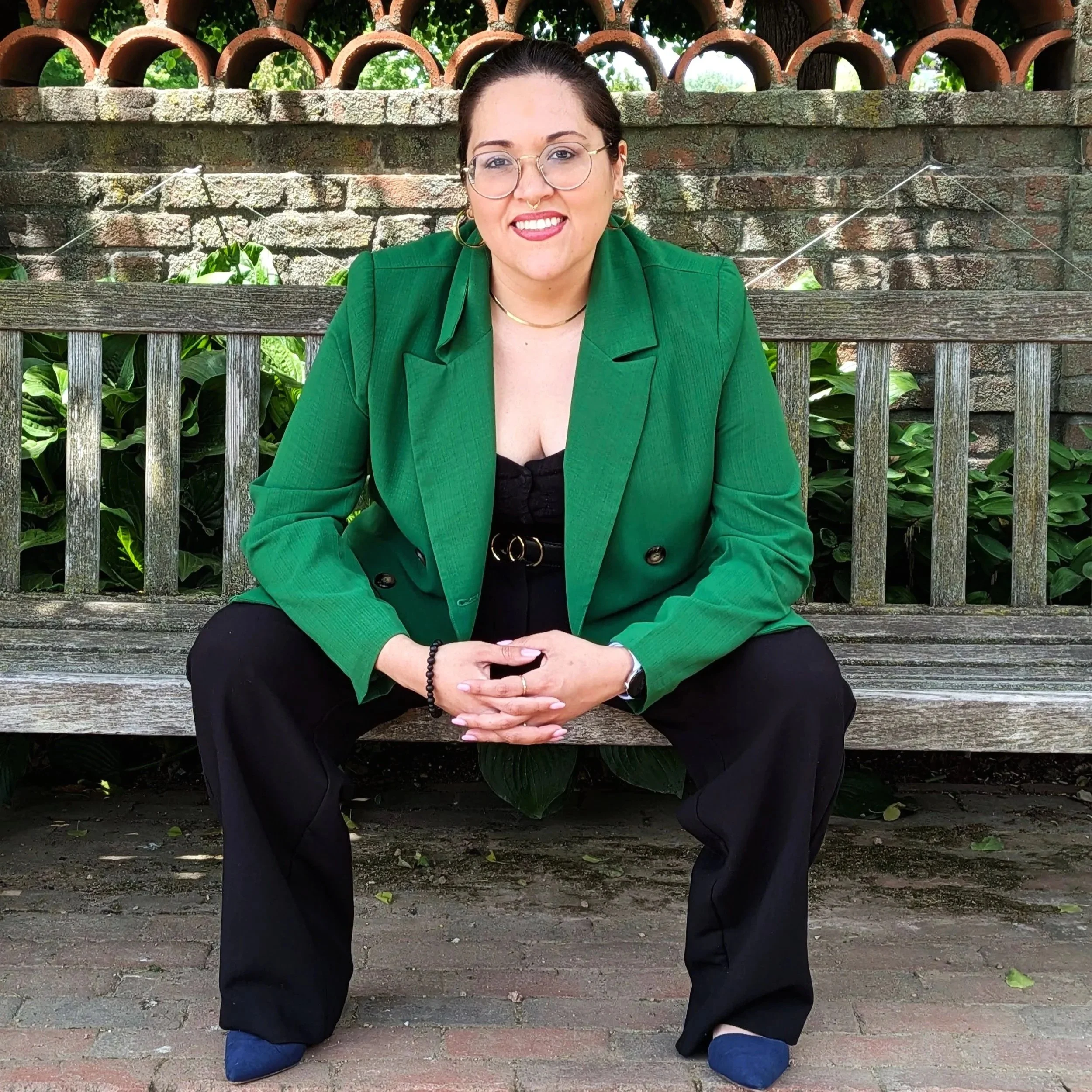 A woman with glasses wearing a green blazer, black dress, and blue slippers sitting on a wooden bench outdoors in front of a brick and tile wall with greenery behind her.