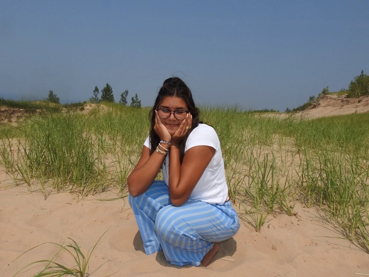 A woman with glasses and dark hair, wearing a white shirt and blue striped pants, sitting on sandy ground with green grass behind her, smiling with eyes closed and hands on her cheeks, outdoors in a natural setting.