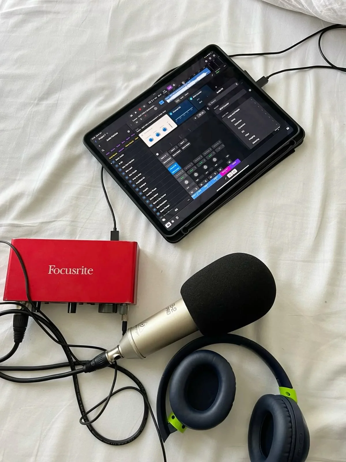 A tablet displaying music production software, connected to a red Focusrite audio interface, with a microphone and headphones nearby on a white surface.