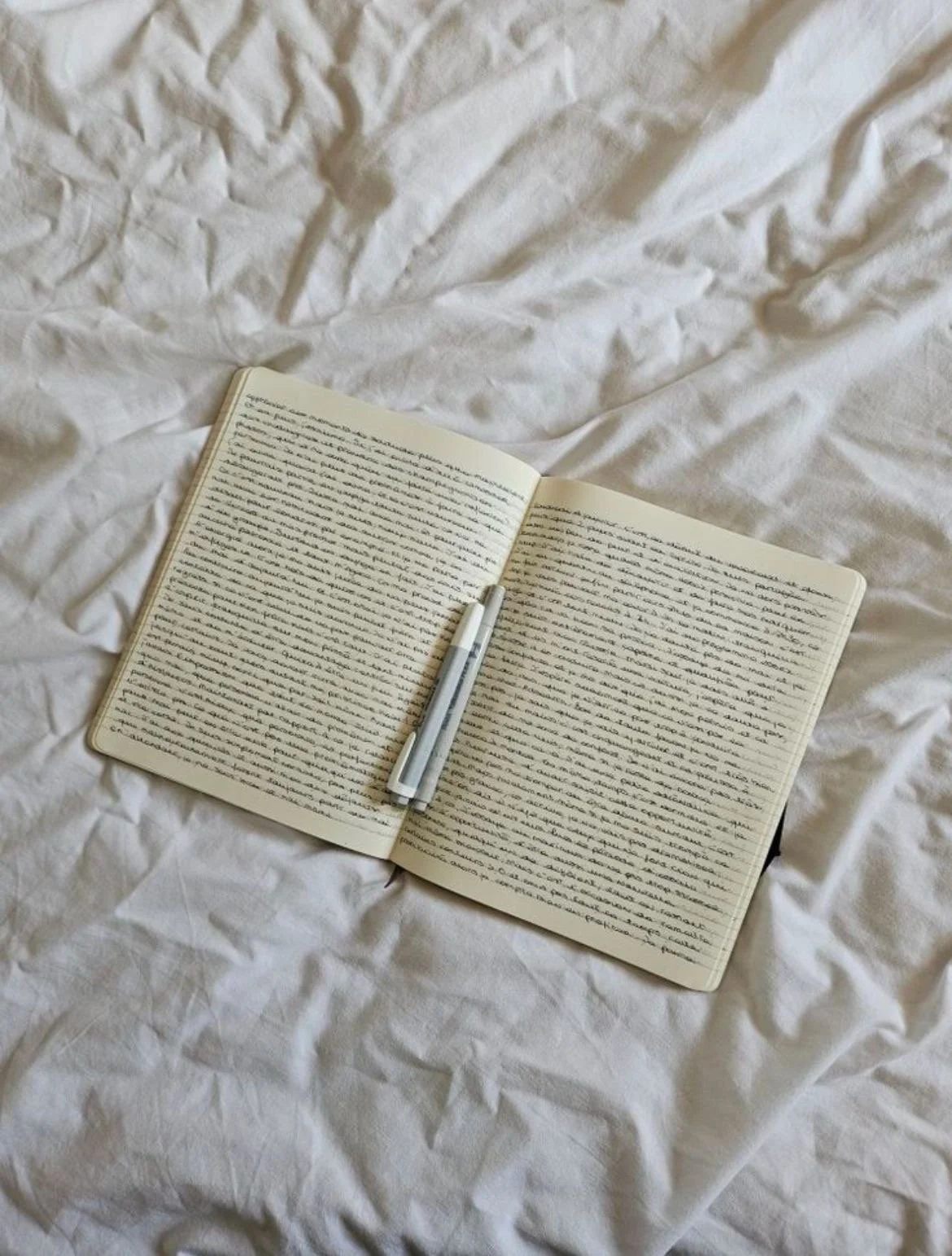 Open notebook with handwritten text on a crumpled white bed, with two pens placed in the center.
