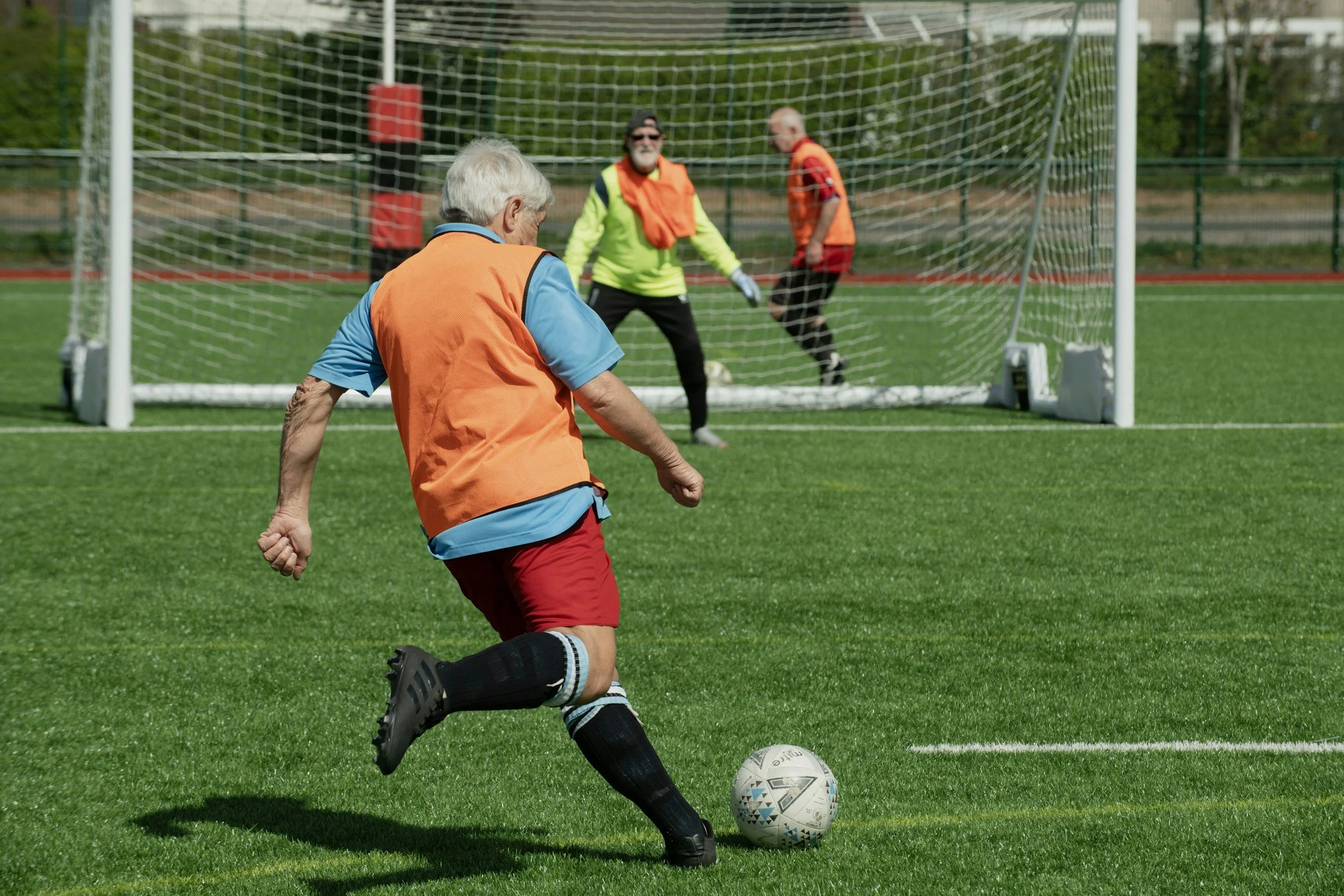 An elderly man playing soccer on a grass field with a goalpost in the background. He is kicking a soccer ball towards the goal, where two goalkeepers wearing green and orange jerseys are preparing to block the shot.