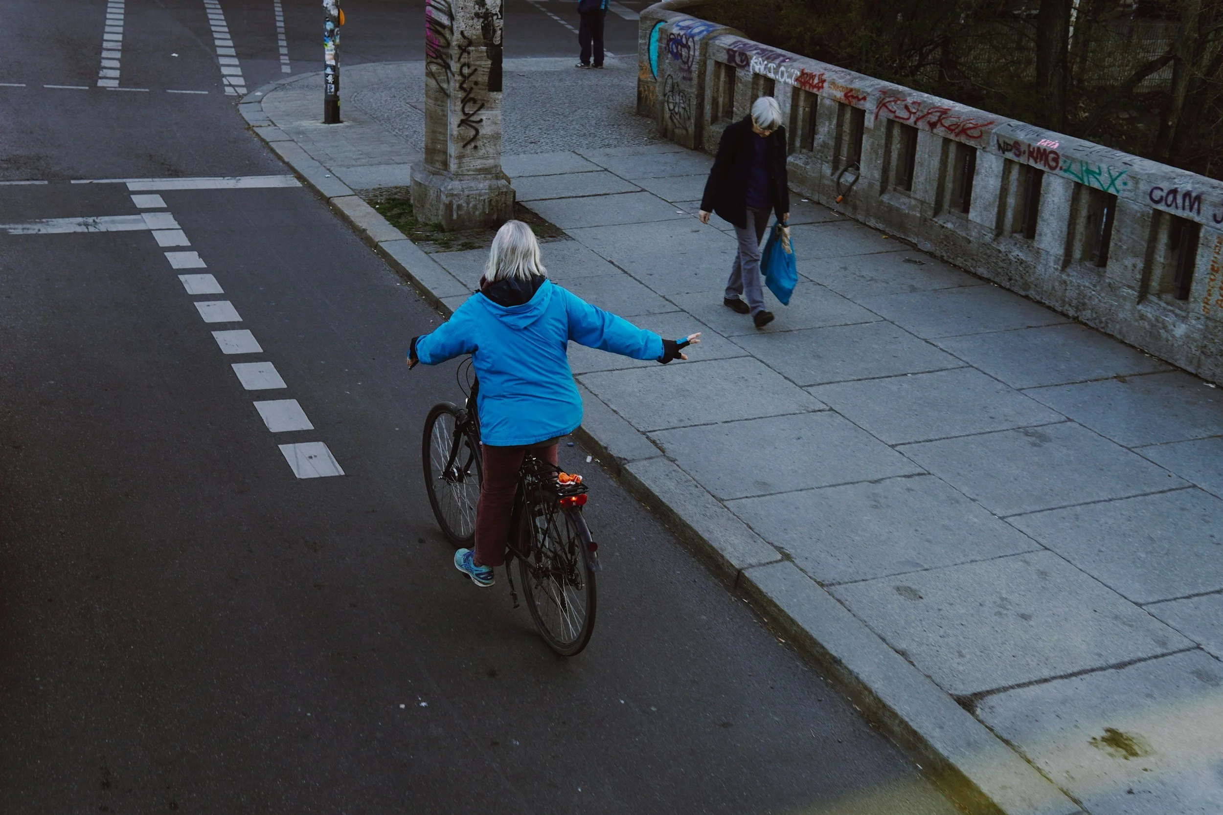 A person riding a bicycle on a city street near the sidewalk, while another person walks on the sidewalk carrying a blue bag, with graffiti-covered wall in the background.