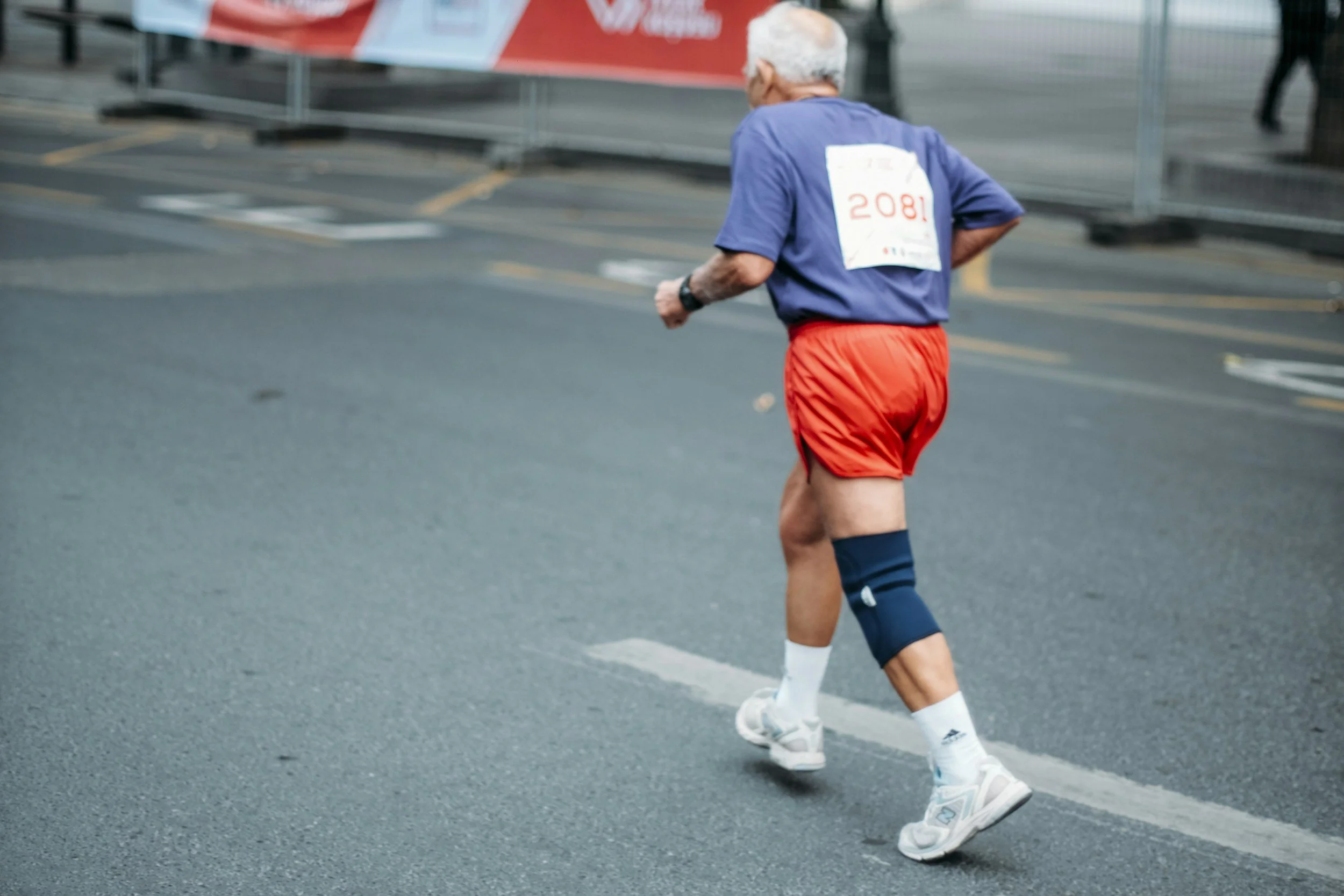 An elderly man running in a race, wearing a blue shirt, red shorts, white athletic shoes, and a knee brace on his right knee. He has a race bib with the number '2081' on his back.