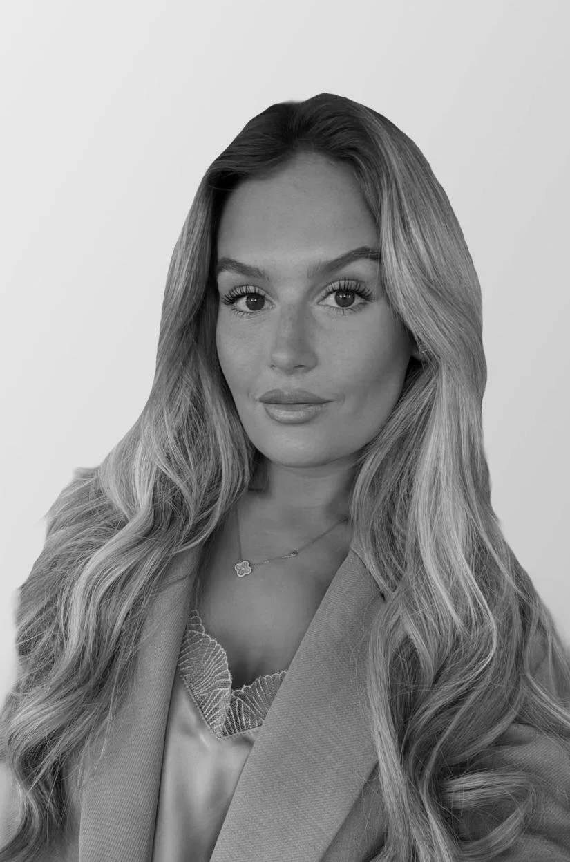 Black-and-white portrait of a woman with long wavy hair, wearing a blazer and a necklace, looking directly at the camera.