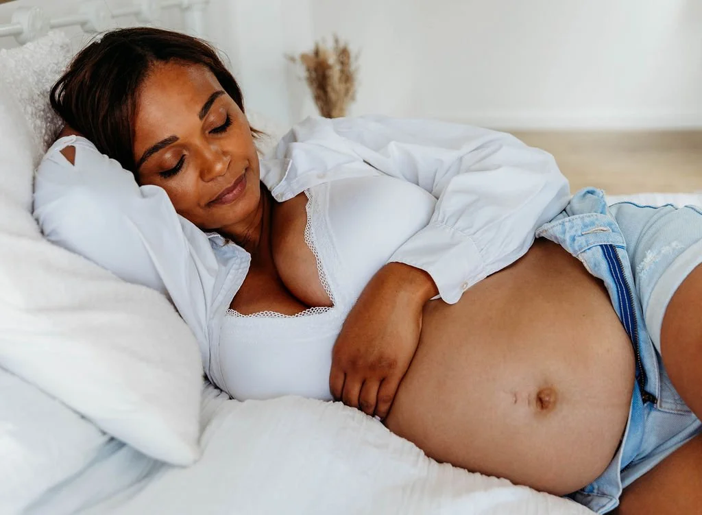 A pregnant woman lying on her side on a bed, resting with her eyes closed, wearing a white top and denim shorts.