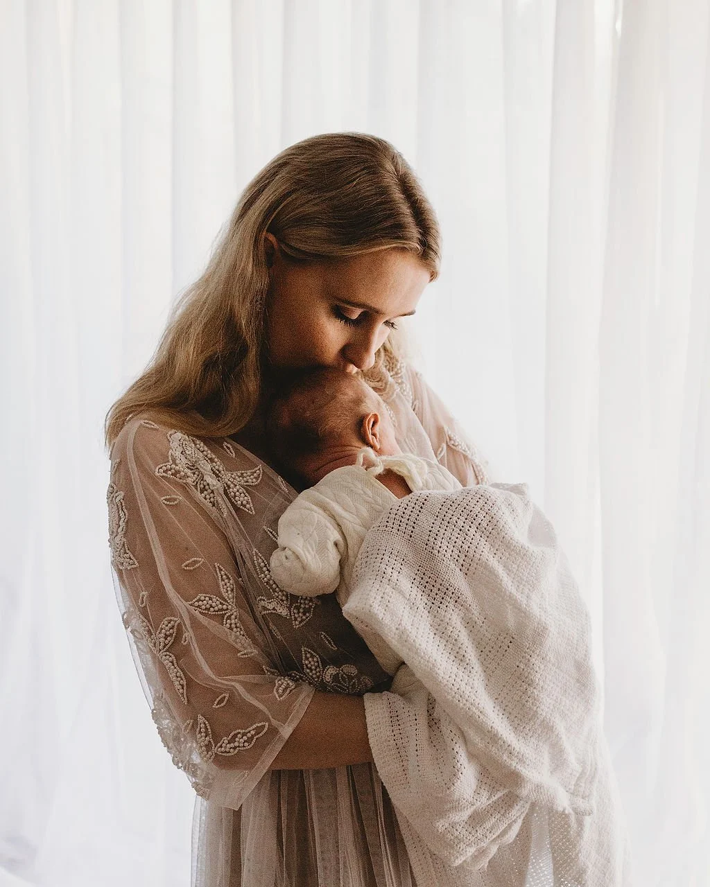 A woman with light brown hair holds a newborn baby wrapped in a white blanket, leaning her forehead against the baby's head in front of a sheer white curtain.