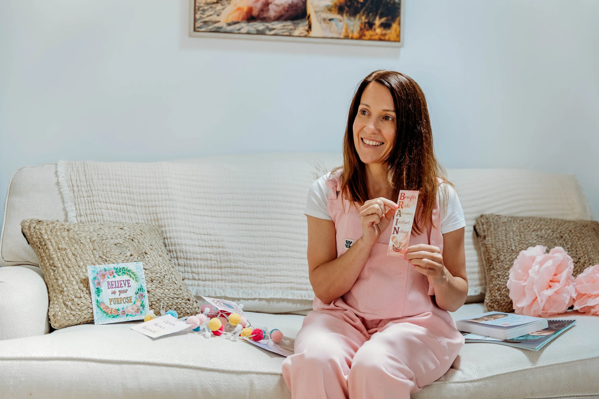 A woman sitting on a cream-colored sofa holding a bookmark with inspirational words, surrounded by colorful decorations and books.