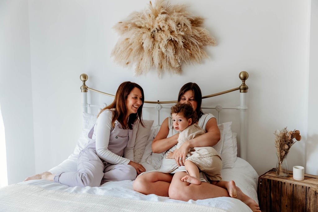 Three women, one holding a young child, sitting on a bed in a bedroom with white walls, a decorative pampas grass wall hanging, and a wooden bedside table with a vase of dried flowers and a candle.