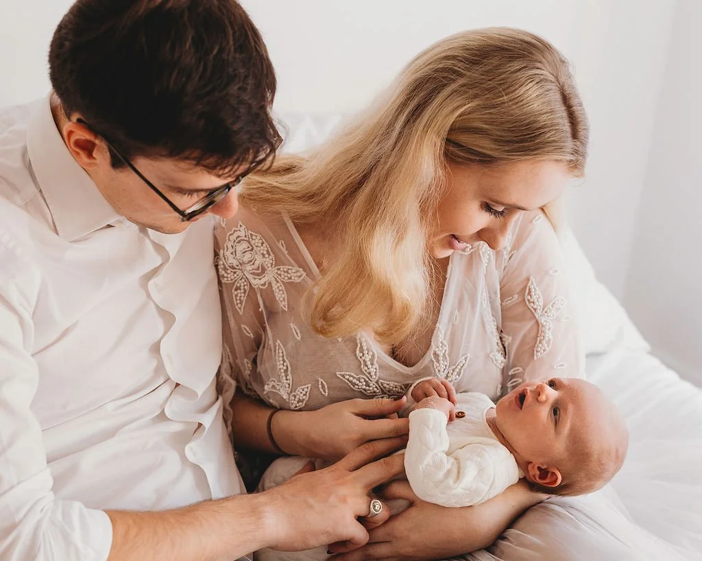 A baby lying on a woman's lap, with a man leaning over and touching the baby's belly, all smiling and in a cozy indoor setting.