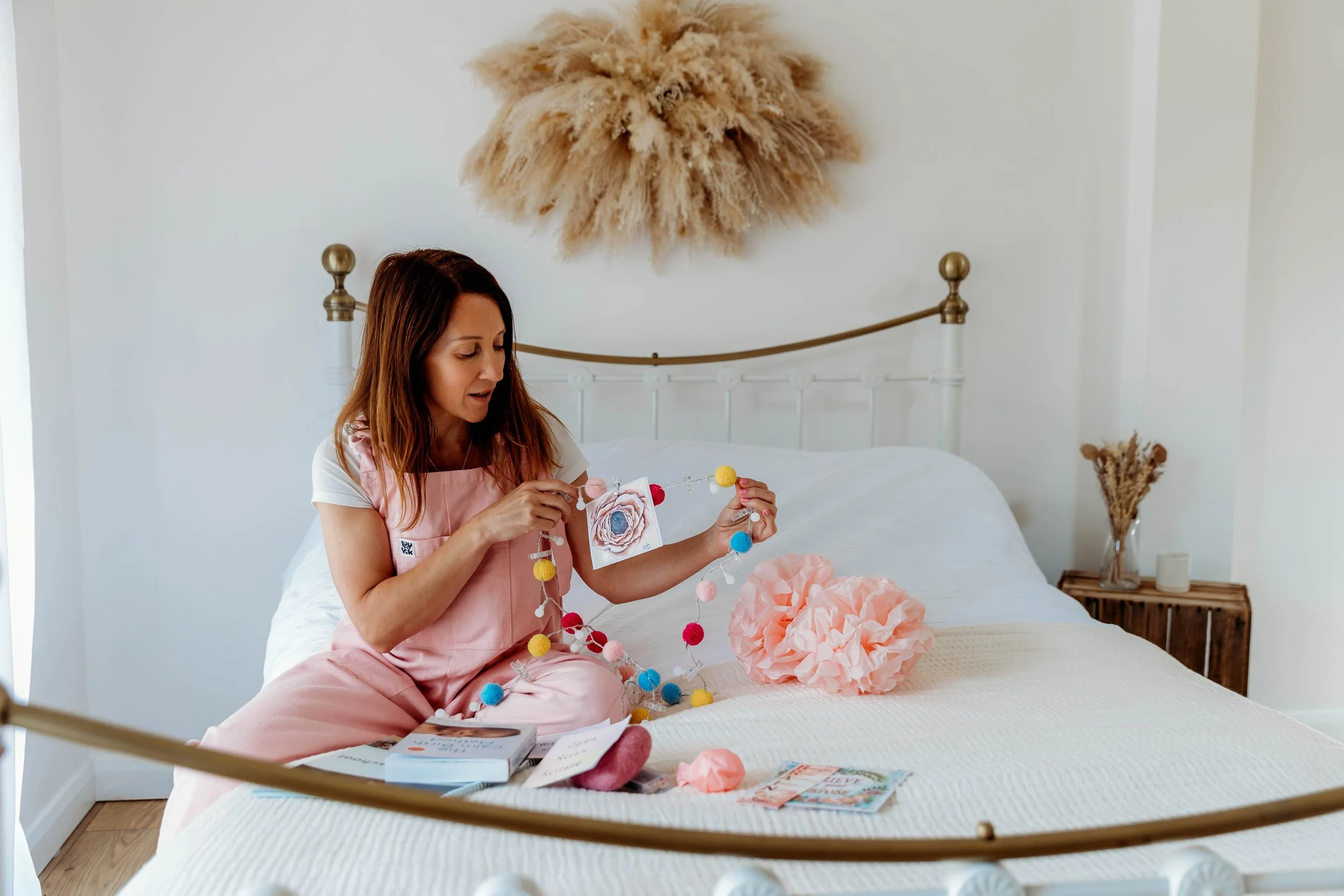 A woman sitting on a bed, decorating a garland with colorful felt balls in a cozy bedroom with white walls, a brass bedframe, and a decorative pampas grass wall hanging.