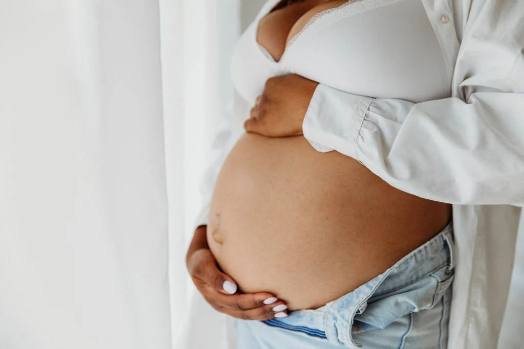 Close-up of a pregnant woman holding her belly, wearing a white shirt and denim shorts.