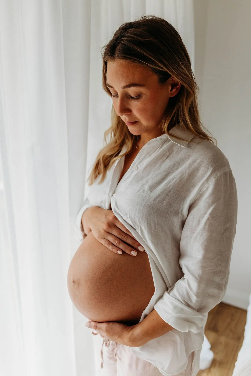 A pregnant woman gently touching her baby bump, wearing an open white shirt, standing near a window with white curtains.